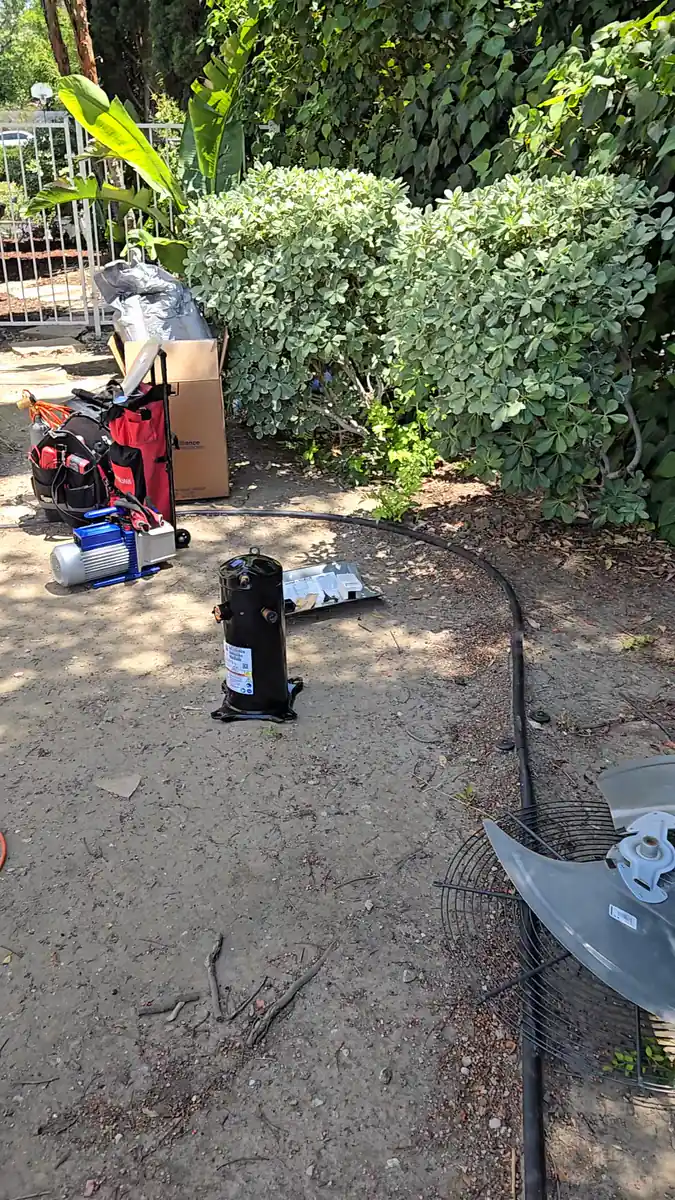 Un compresor de aire acondicionado quemado parado en la tierra del patio de una casa, con el aspa y la rejilla del ventilador del condensador tiradas al lado
