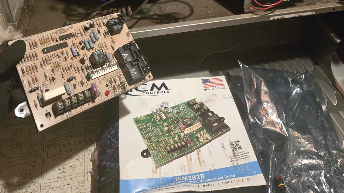 A furnace control board held in a gloved hand in front of an open furnace, with a replacement control board's box on the floor