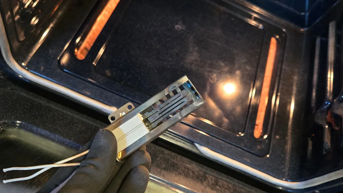Flat gas oven igniter in a slotted metal guard, held in a gloved hand in front of an open oven
