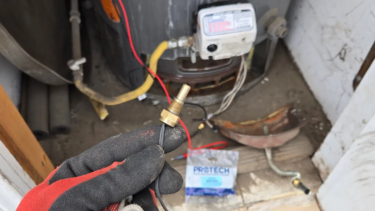 Mano con guante sosteniendo un pequeño sensor de cámara de latón con cable negro frente a la válvula de control de un calentador a gas, con una bolsa ProTech en el piso