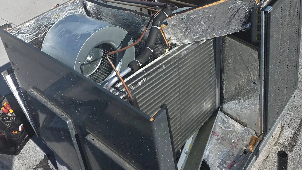 Goodman package unit on a flat roof with its top and panels off, showing the blower and the indoor coil with its foil-faced insulation pulled back