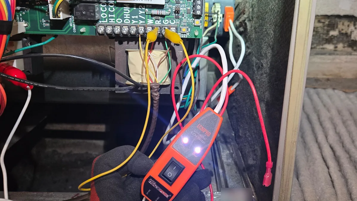 An orange universal ECM tester in a gloved hand, clipped to the terminal strip of the control board inside a Lennox furnace, with both indicator lights on