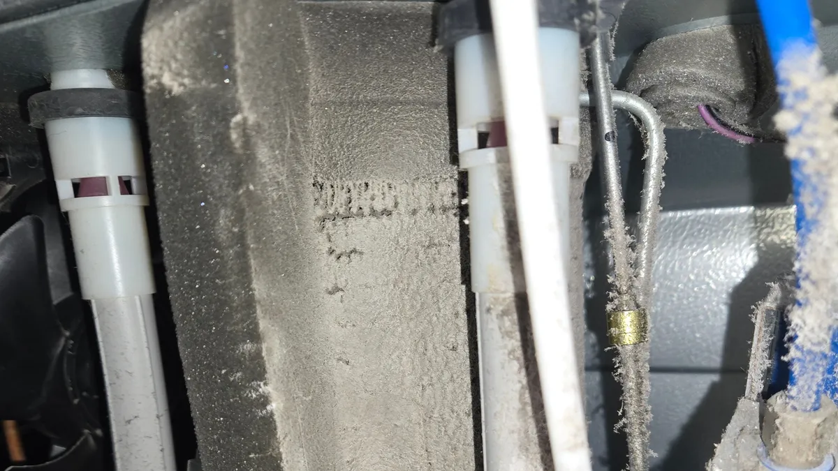 The condenser compartment of a Samsung refrigerator before cleaning, its foam shroud and tubing under a thick gray layer of dust, with lint hanging from a water line