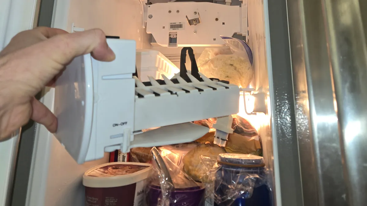 Máquina de hielo sostenida frente al congelador abierto de un refrigerador Whirlpool de puertas lado a lado, con comida todavía en las repisas