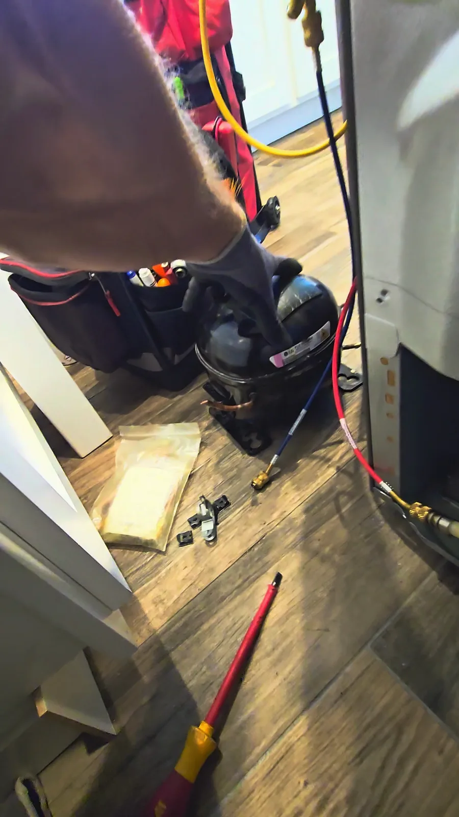 Failed compressor sitting on the kitchen floor next to the refrigerator after removal, with a gloved hand on top