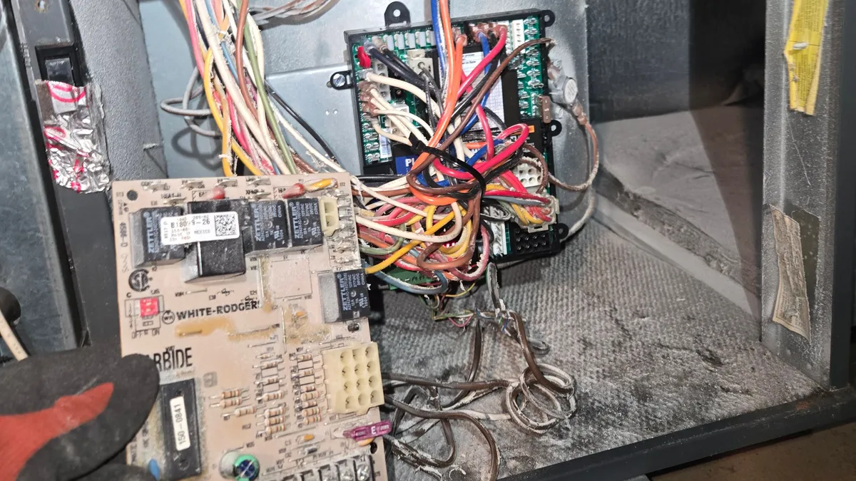 Old furnace control board held in a gloved hand in front of an open Goodman furnace, with the new board wired in behind it